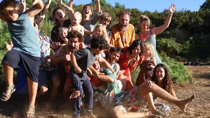 Participants of a La Maison Passerelle gathering, adults and children huddled together in the grass, arms up and laughing.